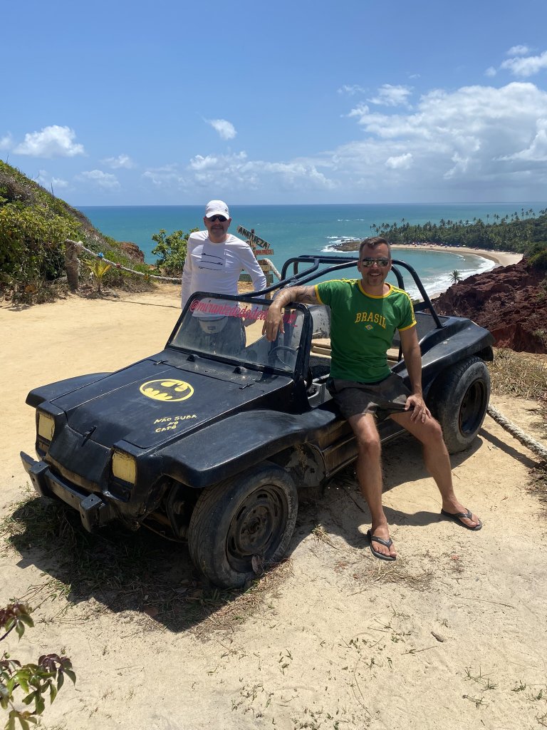 Gringo und Arne sitzen auf einem schwarzen Buggy im Batman-Stil am Aussichtspunkt Dedo de Deus, im Hintergrund Palmen, Strand und das Meer von Tabatinga.