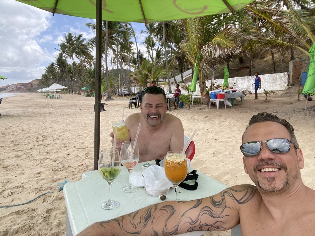 Gringo und Arne sitzen am Tisch mit Cocktails am Strand von Coqueirinho, im Hintergrund grüne Sonnenschirme und Palmen – der Moment vor der Cashew-Geschichte.