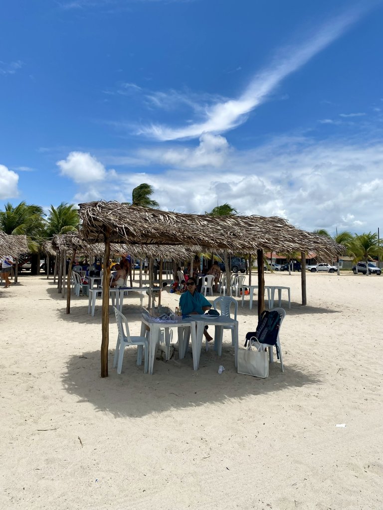 Frau sitzt unter einem Strohdach aus Palmenblättern an einem Tisch am breiten Sandstrand des Praia de Lucena, im Hintergrund weitere Strohdächer und Palmen unter blauem Himmel