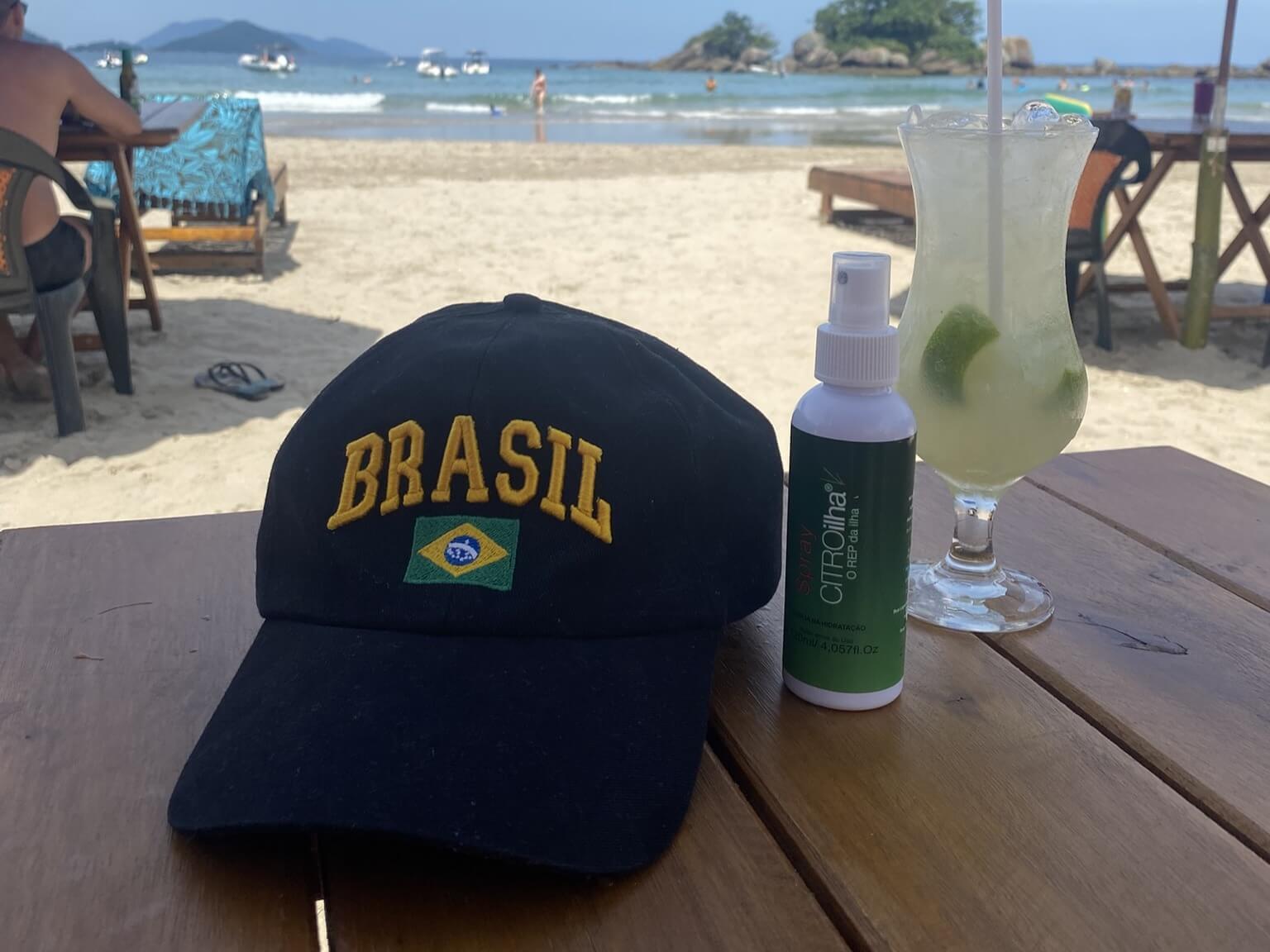 Basecap mit Brasilien-Flagge, Anti-Moskito-Spray und Caipirinha am Strand in Brasilien