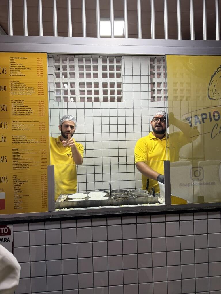 Zwei Köche bereiten Tapioca an einem Straßenstand in Brasilien zu und blicken durch das Fenster